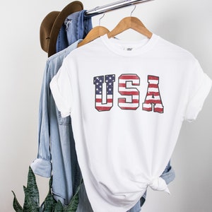 May include: White t-shirt with a distressed American flag graphic that spells out "USA" in large letters.