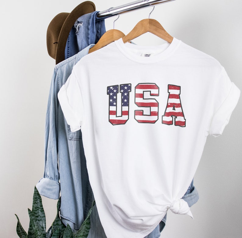 May include: White t-shirt with a distressed American flag graphic that spells out "USA" in large letters.
