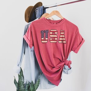 May include: A red t-shirt with a vintage-style graphic of the American flag and the word "USA" printed on the front.