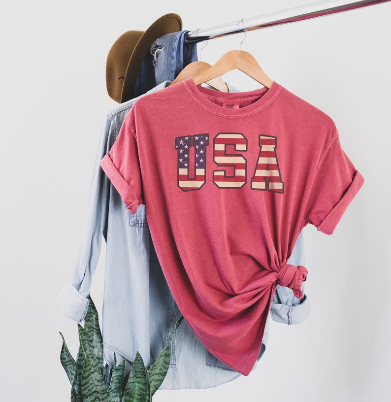 May include: A red t-shirt with a vintage-style graphic of the American flag and the word "USA" printed on the front.