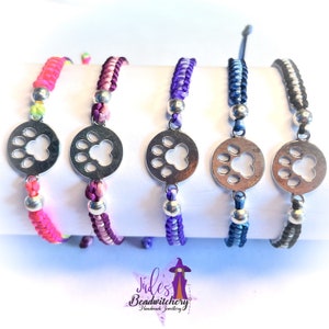 May include: Five colourful friendship bracelets with silver paw print charms. The bracelets are made with a variety of colours, including pink, purple, blue, and black.
