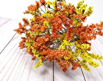 May include: A vibrant bouquet of dried flowers in shades of yellow and orange. The flowers are arranged in a dense cluster, with a mix of textures and shapes. The background is a light-colored wooden surface.