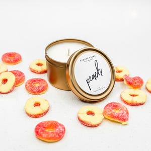 May include: A gold-colored candle tin with a white label that reads "peach" in script. The candle is surrounded by peach-shaped gummy candies. The label also includes the text "ALWAYS EXTRA" and "hand-poured soy candle".