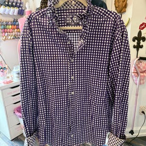May include: A purple and white gingham patterned shirt with a ruffled collar and cuffs. The long-sleeved shirt is button-down and hangs on a pink clothing rack. The shirt has a small emblem on the chest.
