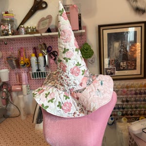 May include: A quilted pointed hat featuring a white base with a floral pattern of pink roses and green leaves. The hat has a wide, flexible brim and a tall, conical crown, constructed from fabric with visible stitching throughout the material.