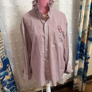 May include: A long-sleeved button-down shirt featuring a small red and white gingham check pattern. The shirt has a high ruffled collar and a red embroidered OU logo on the left chest pocket area. It is displayed on a white mannequin torso.