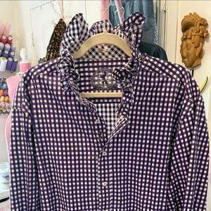May include: A long-sleeve button-down shirt featuring a purple and white gingham check pattern. The shirt includes a prominent ruffled collar, a front button placket with white buttons, and an interior label displaying the brand name Eton.