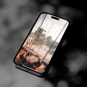 Può includere: Uno smartphone nero mostra una vista panoramica di un edificio con facciata in vetro, con un orologio che segna le 19:02. Il telefono è inclinato su uno sfondo nero e grigio sfocato.