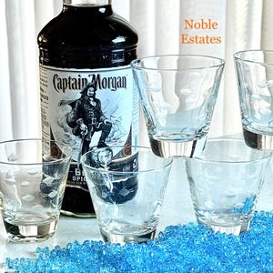May include: A bottle of Captain Morgan spiced rum next to five clear, etched glasses with a fish design. The glasses have silver-colored bases. The background features blue decorative beads and the text "Noble Estates".