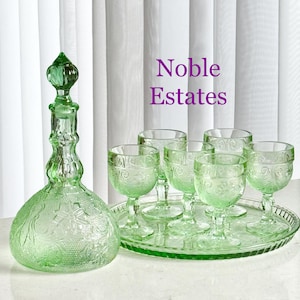 May include: A green glass decanter with a stopper and a set of six matching wine glasses on a tray. The glassware features an embossed floral design. The text "Noble Estates" is visible in the background.