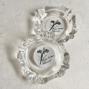 May include: Two clear glass ashtrays with a white circle design featuring a black and white illustration of a ring-necked pheasant. The text "Ring Necked Pheasant" is printed in black on the white circle.