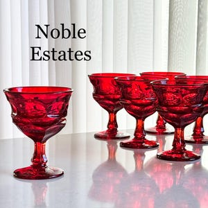 May include: A set of six ruby red vintage stemmed glasses. The glasses have a decorative pattern and are arranged on a reflective surface. The text "Noble Estates" is displayed in black at the top of the image.