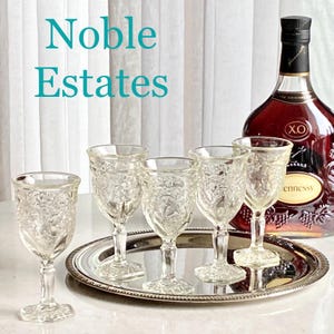 May include: Set of six clear glass stemmed wine glasses with a cut glass design. The glasses are arranged on a silver tray with a bottle of Hennessy XO cognac in the background. The text "Noble Estates" is displayed in the top left corner of the image.