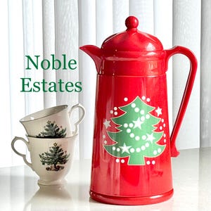 May include: A red Christmas-themed coffee carafe with a green Christmas tree design and two matching teacups. The teacups and carafe have a white background. The text "Noble Estates" is visible.