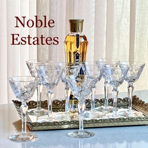 Puede incluir: Conjunto de siete vasos de cóctel de vidrio transparente con diseños florales grabados, dispuestos en una bandeja dorada con una botella de licor en el centro. Los vasos son todos del mismo tamaño y forma, y la botella es alta y esbelta. El texto "Noble Estates" está escrito en letras rojas en la parte superior de la imagen.