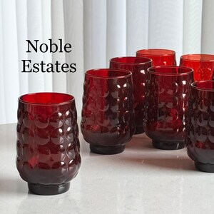 May include: A collection of ruby red drinking glasses with a textured, bubble-like pattern. The glasses have a dark base and are arranged on a light surface. The text "Noble Estates" is visible in the image.