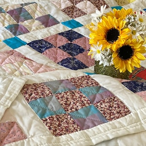 May include: A white patchwork quilt featuring a variety of floral patterns and colours, including pink, blue, and yellow. A bouquet of sunflowers and white daisies is placed on top of the quilt.