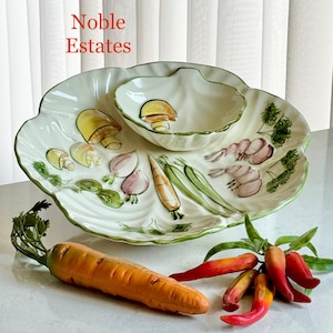 May include: A white ceramic serving platter with a scalloped edge and a smaller bowl, both decorated with painted vegetables. The platter features mushrooms, garlic, carrots, and peas. A large orange carrot and red peppers are in the foreground. The text "Noble Estates" is visible.