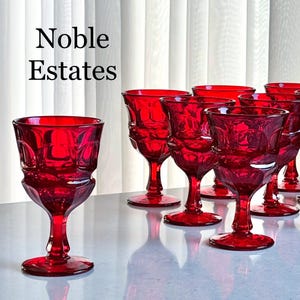 May include: A set of ruby red wine glasses with a detailed, geometric design. The glasses are arranged on a reflective surface, with the text "Noble Estates" in the upper left corner. The stemware is ideal for serving wine.