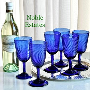May include: A collection of six cobalt blue wine glasses with a spiral design, arranged on a silver tray. A bottle of white wine stands to the left. The text "Noble Estates" is in green.