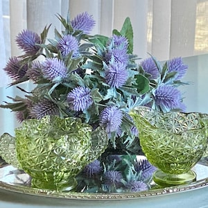 May include: Two green glass bowls with ruffled edges and a starburst pattern sit on a silver tray. A bouquet of purple thistle flowers is in the background.