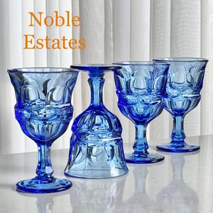May include: Four cobalt blue wine glasses with ornate, textured designs. The glasses are displayed on a reflective surface, with the text "Noble Estates" in the upper left corner. The glasses have a classic, elegant shape.