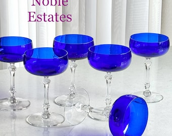 May include: Set of six cobalt blue champagne coupe glasses. The glasses have a round bowl, a long stem, and a circular base. One glass is lying on its side. The text "Noble Estates" is in the upper left corner.
