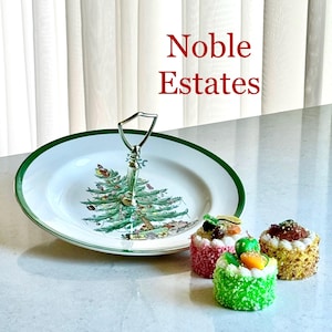 May include: A two-tiered serving tray with a green rim and a Christmas tree design. The tray has a silver handle and is accompanied by three small, colorful, decorative cakes. The text "Noble Estates" is displayed in red.