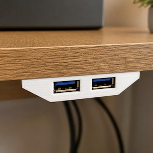 Puede incluir: Un concentrador USB blanco de dos puertos montado en la parte inferior de una superficie de escritorio de madera. El dispositivo cuenta con dos puertos rectangulares con componentes internos de plástico azul para cables y periféricos.