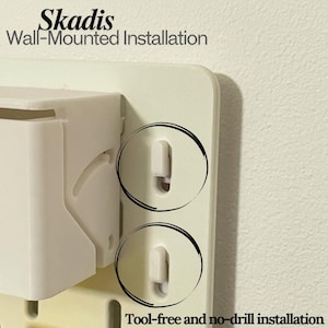 Può includere: Primo piano di un sistema di installazione a parete color crema. Il sistema presenta un componente rettangolare bianco con due ritagli circolari che evidenziano l'installazione senza attrezzi e senza foratura. Il testo "Skadis Wall-Mounted Installation" è visibile.