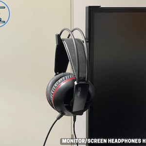 May include: Black and red headphones with the brand name "ASUS" are hanging on a black monitor screen holder. The holder is attached to the monitor. The text "MONITOR/SCREEN HEADPHONES HOLDER" is at the bottom.