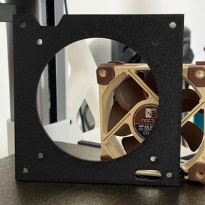 May include: A black square metal fan mount with a circular opening, showcasing a beige and brown computer fan. The fan has brown blades and a beige frame. The mount has four screw holes. A second fan is visible.