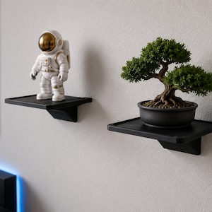 May include: Two black wall-mounted shelves with a square surface and a support bracket underneath. One shelf holds a white astronaut figurine with a gold visor, and the other shelf holds a small green bonsai tree in a black circular pot.