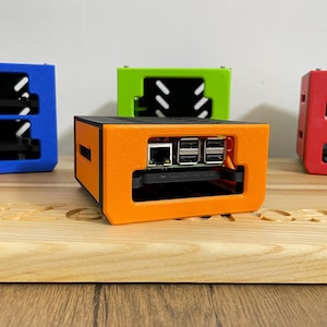 Op de afbeelding: Een oranje en zwarte Raspberry Pi-behuizing met zichtbare poorten, geplaatst op een houten oppervlak. Andere behuizingen in blauw, groen en rood staan op de achtergrond. De behuizingen hebben een rechthoekig ontwerp met ventilatiesleuven.
