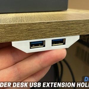 Puede incluir: Un soporte blanco para extensión USB debajo del escritorio con dos puertos USB. El soporte está montado debajo de un escritorio de madera. Los puertos USB tienen inserciones azules. El texto "DUAL UNDER DESK USB EXTENSION HOLDER" es visible.