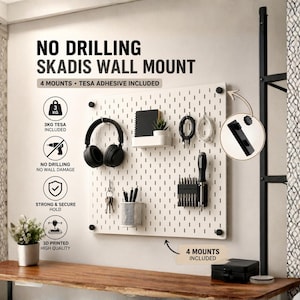 May include: A white rectangular pegboard wall mount with black corner fasteners. Text reads NO DRILLING SKADIS WALL MOUNT. Accessories include a white shelf, a metal mesh cup, and black hooks holding headphones, keys, cables, and tools. Includes 4 mounts and Tesa adhesive for a 3kg load capacity.