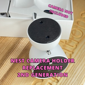 Op de afbeelding: Witte Nest camera houder vervanging voor de 2e generatie. De houder heeft een zwarte, cirkelvormige bovenkant en een witte basis. De tekst "CAMERA NOT INCLUDED" is roze. De houder staat op een houten oppervlak.