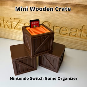 May include: Miniature brown wooden crate with an orange interior, holding a Nintendo Switch game cartridge. The crate is part of a stack of three, with the text "Mini Wooden Crate" and "Nintendo Switch Game Organizer" visible.