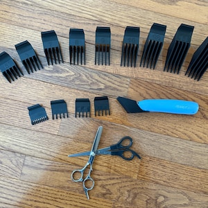 Puede incluir: Un conjunto de protectores negros para cortapelos, una maquinilla de afeitar azul y negra y unas tijeras plateadas y negras están dispuestas sobre una superficie de madera. Los protectores están numerados del 1/8 al 16.