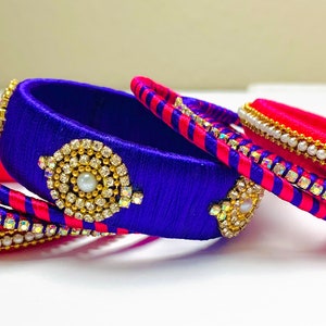 May include: A set of six silk thread bangles, two are pink with gold and white bead accents, and four are blue with gold and white bead accents. One blue bangle has a large rhinestone embellishment.