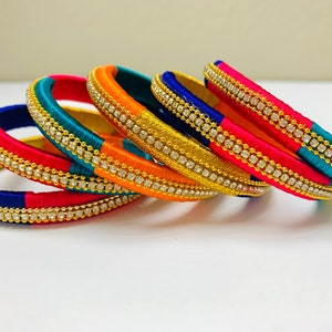 May include: A set of six colorful silk thread bangles with gold accents and clear rhinestones. The bangles are stacked in a pile, with the blue and pink bangles on the bottom and the orange and turquoise bangles on top.