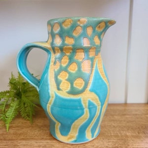 May include: A turquoise ceramic pitcher with a yellow and green abstract design. The pitcher has a wide mouth and a handle.