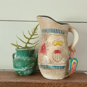May include: A beige ceramic pitcher with a black rim, handle, and colorful geometric designs. The pitcher has a tag that reads "Indian Style Ceramics". A small green vase with a plant is next to the pitcher.
