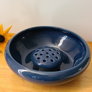 May include: A deep blue ceramic bowl with a smaller, matching blue ceramic flower frog in the center. The bowl has a rounded shape and a smooth, glossy finish.