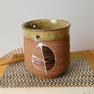 May include: A brown ceramic planter with a leaf design. The planter has a speckled glaze and a hole for drainage. It is hanging from a rope.