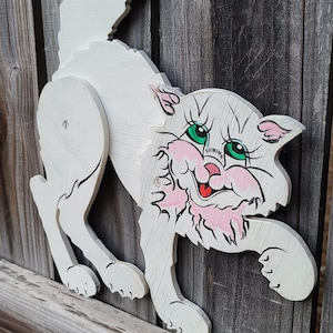 May include: A white wooden cat cutout with a playful expression. The cat has green eyes, pink cheeks, and a red tongue. It is painted on a wooden plank and appears to be mounted on a wooden fence.