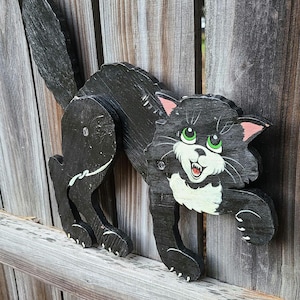 May include: A black wooden cat decoration with a playful design. The cat has green eyes, pink ears, and a white chest. It is mounted on a wooden fence, creating a whimsical outdoor accent. The cat's tail is raised, and it appears to be in a playful pose.