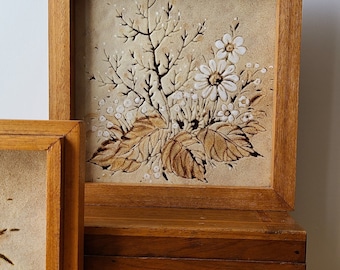 May include: A framed ceramic tile with a floral design. The tile is beige with brown and white flowers and leaves. The frame is made of wood and is a light brown color.