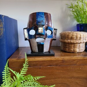 May include: A decorative wooden mask with inlaid blue, brown, and white stones. The mask has a stylized face with large eyes and a wide mouth. The mask is sitting on a wooden shelf with a fern plant in the foreground.