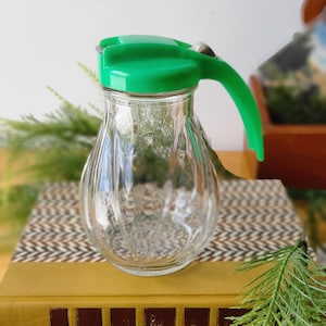 May include: Clear glass syrup dispenser with a green lid and handle. The dispenser is sitting on top of a stack of books. The books are brown and green.